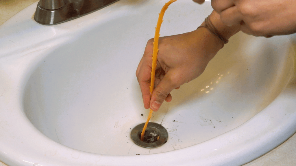 The image shows a man applying a drain snake to fix a clogged bathroom sink.