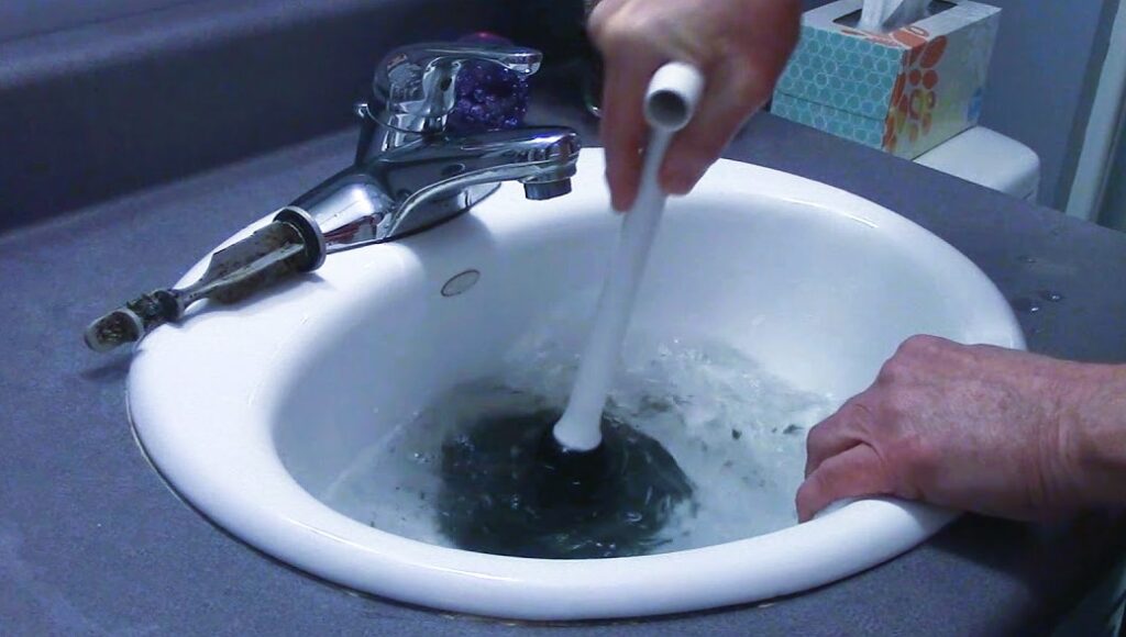 The image shows a man applying a plunger to fix a clogged bathroom sink.