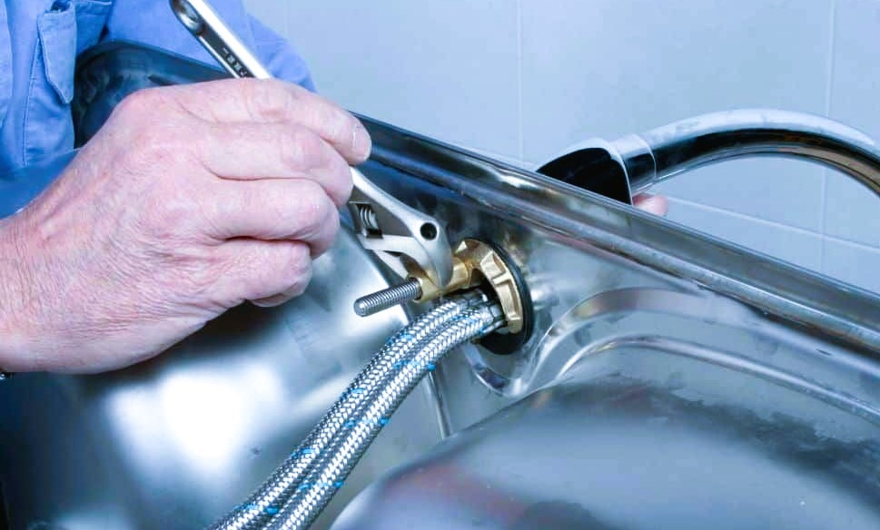 The image shows a man tightening a loose kitchen sink faucet.