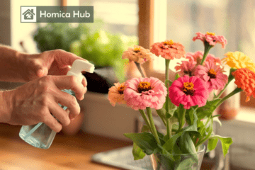 The image shows a man's hands holding a spray bottle in hands to spray on the zinnias, the vase of zinnias is placed on wooden countertop.